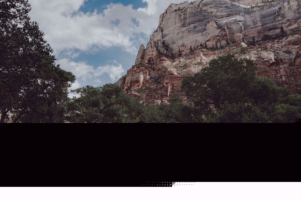 A rustic 1925 stone cabin, nestled around scrub and trees. A rock formation towers in the background.
